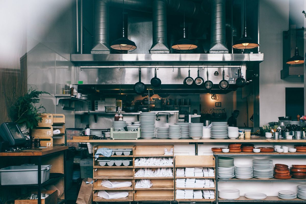 Restaurant kitchen with stainless steel hoods, hanging pans and open shelves stacked with plates and bowls